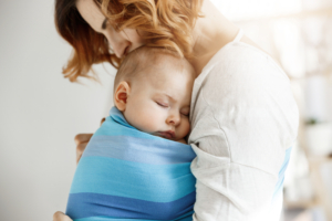 Precious little newborn boy having deep sleep at day on mother chest in blue baby sling. Mom kissing baby head and feeling relaxed and delight. Family concept.